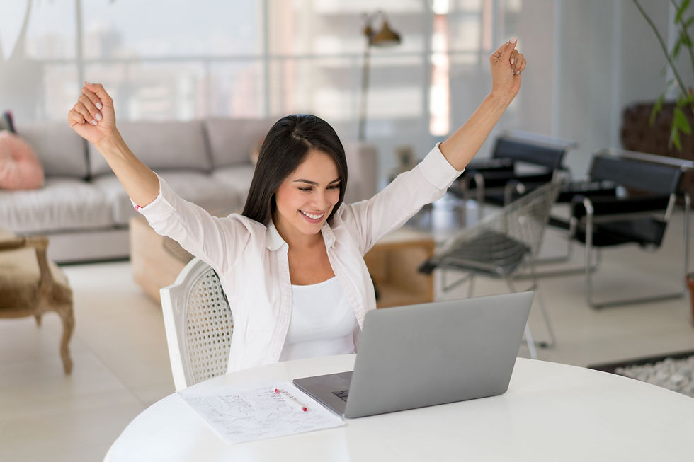 Smiling woman at a laptop in a bright living room, arms raised in celebration, with papers on the table.