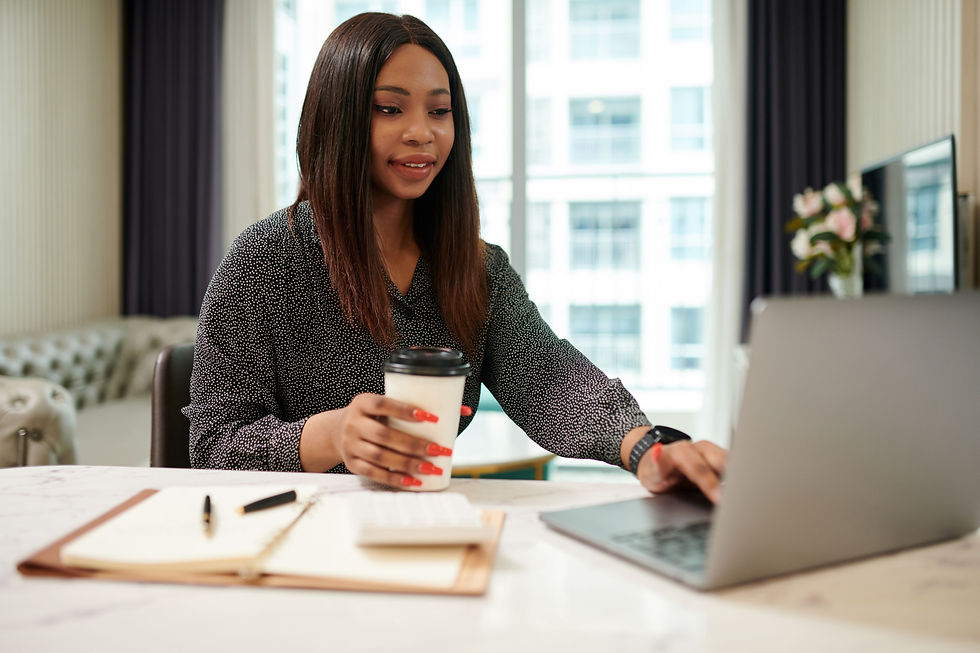 Woman seated at a desk working on a laptop while holding a takeaway coffee, with a notebook and pen on the table.