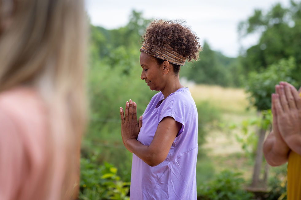 A woman in a purple shirt meditates with hands in prayer position outdoors. Blurred figures and lush greenery create a peaceful vibe.