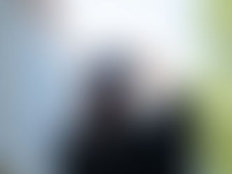 Graduate in a black cap and gown holds their cap with a backdrop of a building and greenery, conveying achievement and celebration.