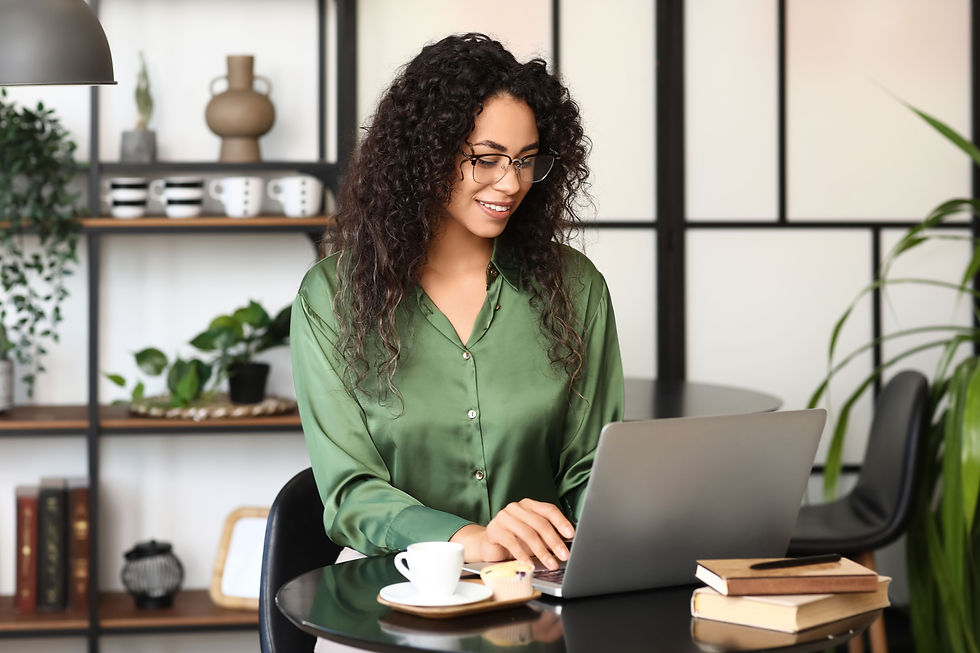 Woman in green blouse using laptop at a round table with books and a coffee cup. Shelves with plants and decor in the background. Calm mood.