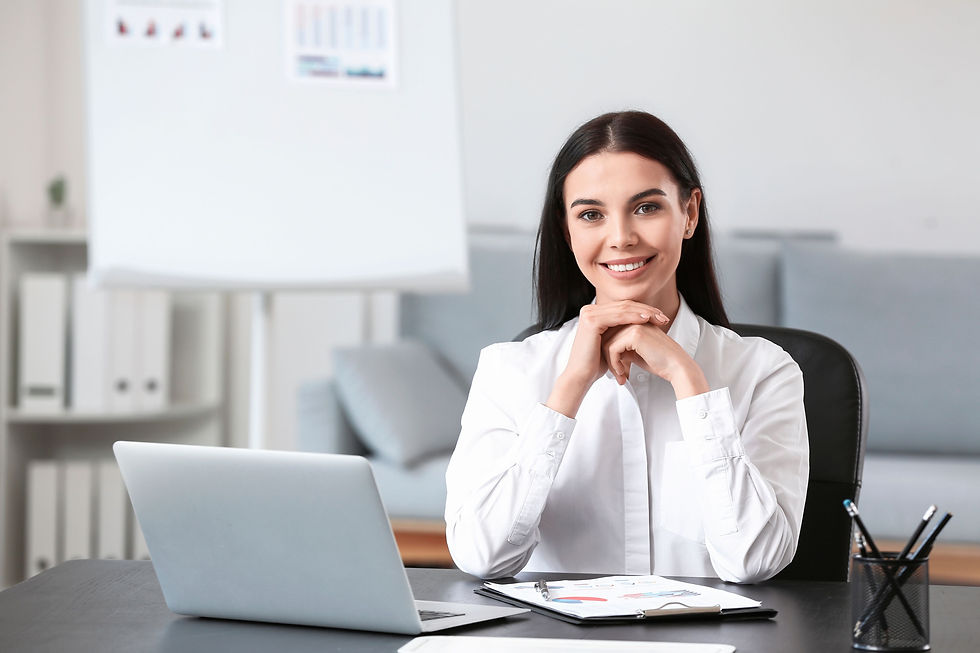 Smiling woman in white shirt seated at office desk with laptop, charts, and pen holder. Gray couch and whiteboard in background.