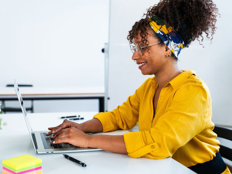 Smiling person in a yellow shirt typing on a laptop at a desk. A colorful headband and bright sticky notes add a cheerful touch.