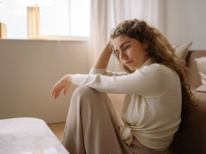 Woman in a cream sweater sits on a beige couch in a sunlit living room, resting her head on her hand and looking pensive.