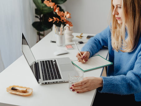 Woman in a blue sweater writes in a notebook at a desk with a laptop. She holds a glass, plants in the background. Bright, focused mood.