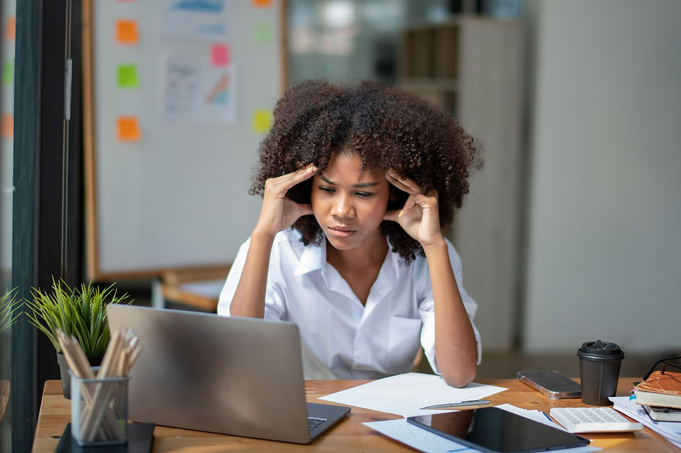 Woman in white shirt looks stressed at laptop, holding her temples. Office setting with papers and coffee cup on desk, colorful sticky notes in background.