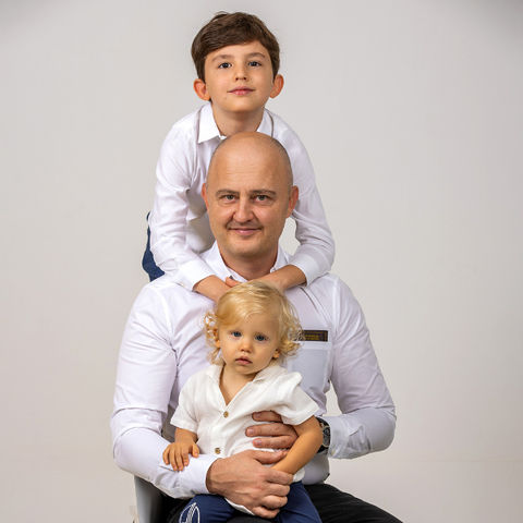 A bald father sitting on a chair with his 9-year-old son hugging him from behind and his 2-year-old daughter on his lap, a beautiful family moment.