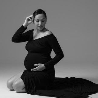 A pregnant woman in a black, long-sleeved gown kneels on the floor in a serene pose, one hand supporting her bump and the other resting softly on her thigh.