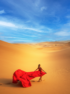 A lady in a red dress standing in the desert, her dress billowing in the wind as she faces the sand dunes.