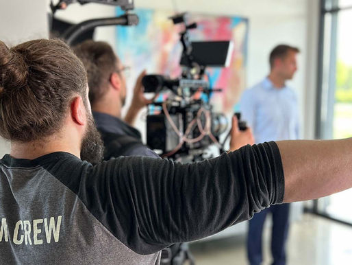 Film crew member points as camera operator films two people in a bright room. Crew's shirt reads "FILM CREW." Blurred colorful painting behind.