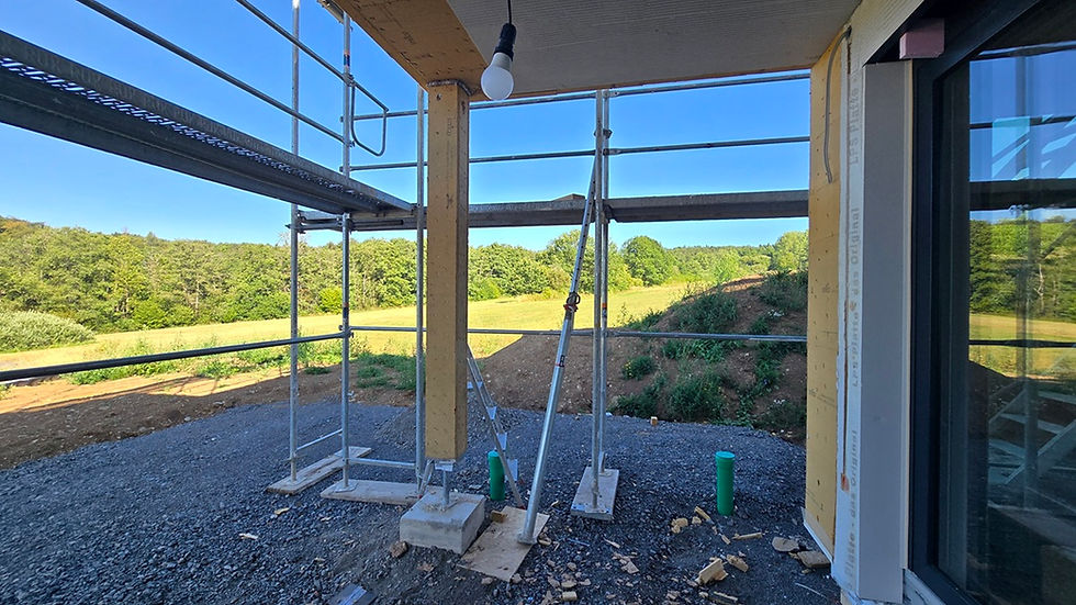 Blick aus dem Haus auf die nun montierte Säule an der Ecke der Terrassenüberdachung. Sie ist mit braunen OSB-Platten verkleidet und hat unten einen dünnen Metallfuß, der auf dem Punktfundament für die Säule verschraubt ist. Der Boden ringsum ist mit Schotter bedeckt, außerhalb der Terrasse sieht man die Metallteile des Gerüsts. Im Hintergrund sieht man hellgrüne Wiesen, etwas dunkler grüne Wälder und darüber blauen Himmel. Im Vordergrund hängt eine Birne an einem Kabel von der Terrassendecke.