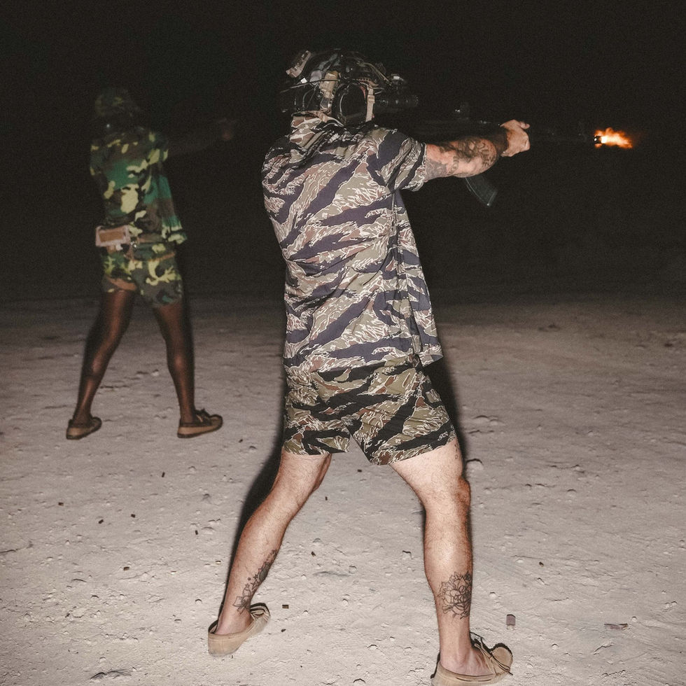 A well lit photo of a person firing an AR15 in the desert and wearing tiger stripe QILO EDC Mk. II shorts