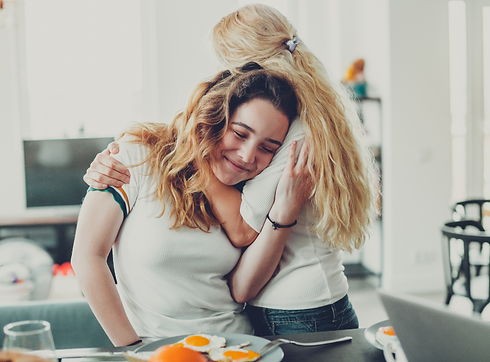 A mother and daughter hug whilst making breakfast. The teenage daughter has her eyes closed and is smiling.
