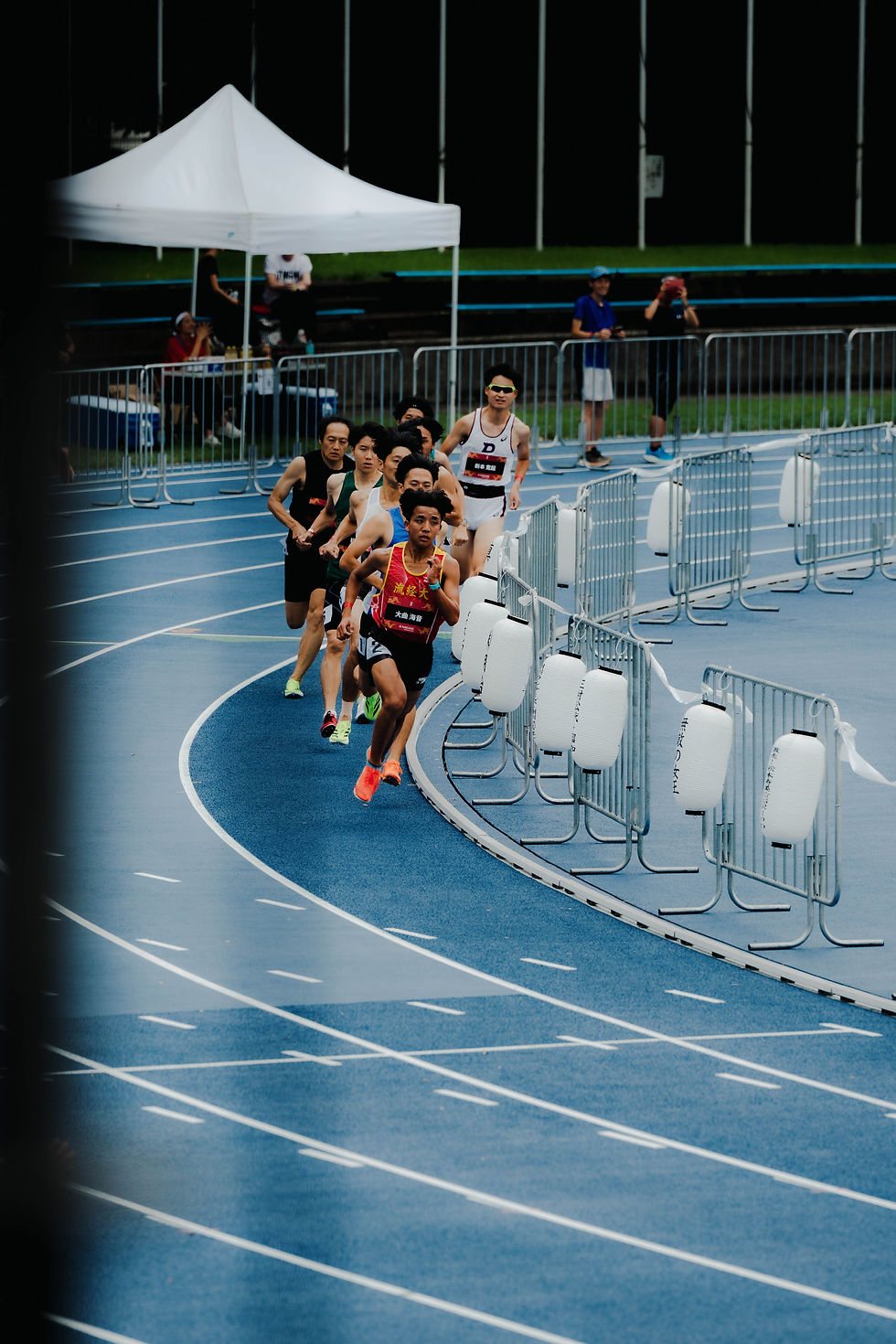 Runners compete on a blue track, rounding a curve with determination. White lanterns line the edge. A white tent is visible in the background.