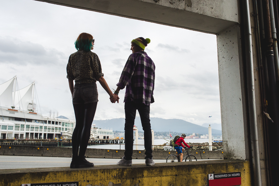 Two people holding hands beneath a concrete beam, with water and a white building behind them.