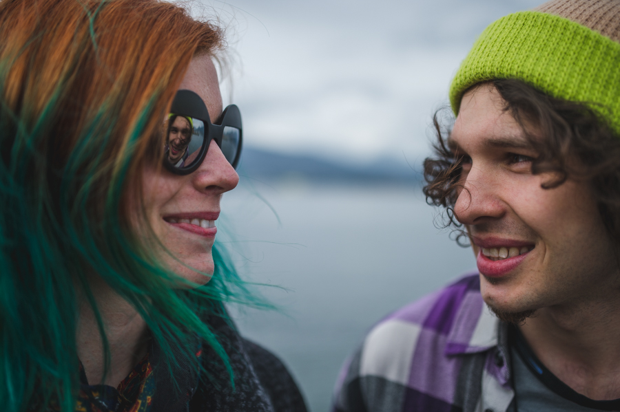 Close portrait of a smiling woman with orange-and-green hair and sunglasses beside a man in a bright knit hat.