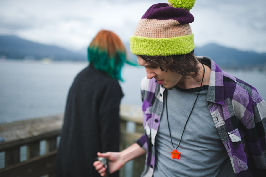A man in a striped knit hat looking down at a wooden toy, with a woman and water behind him.