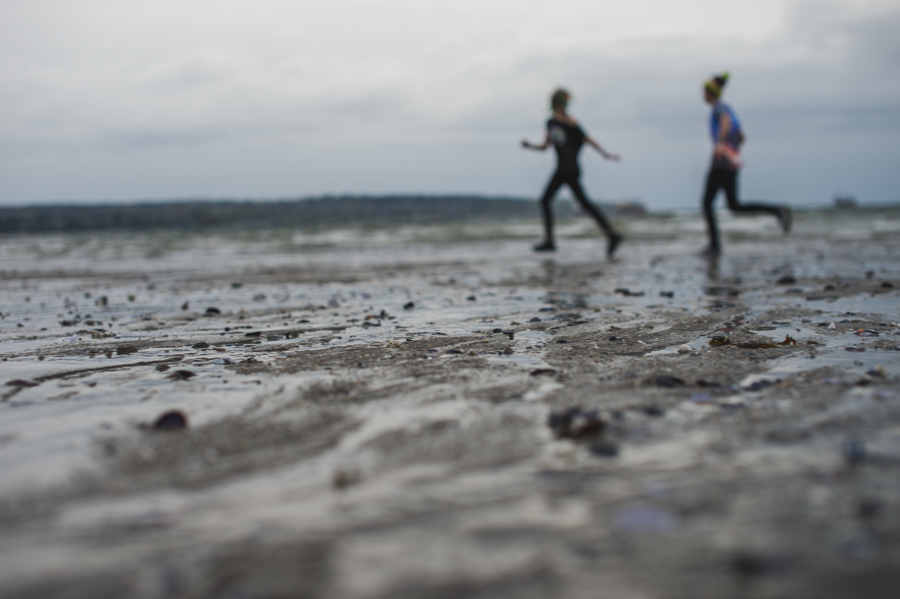 Two out-of-focus people running across a wet beach behind a sharply focused sandy foreground.