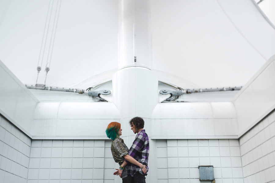 Two people facing each other in a white-tiled stairwell beneath a tall central column.