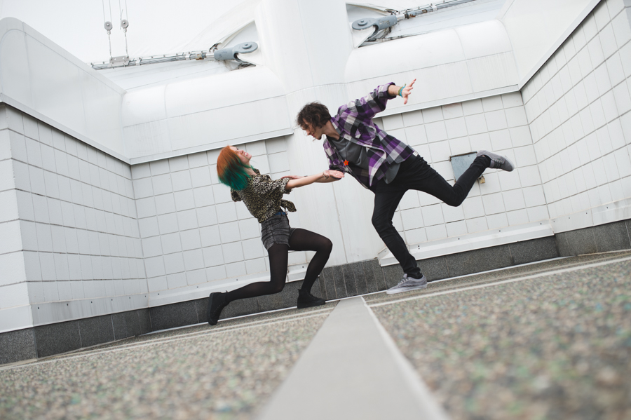 Two people holding hands and lifting their legs in a white-tiled stairwell.