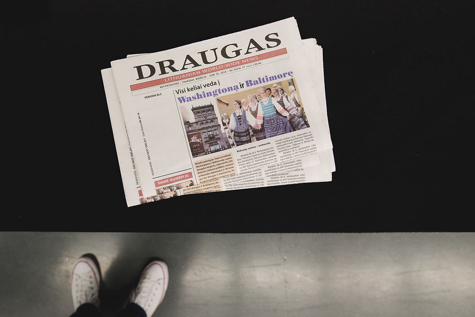 Overhead view of a folded Draugas newspaper above dark flooring and white shoes.
