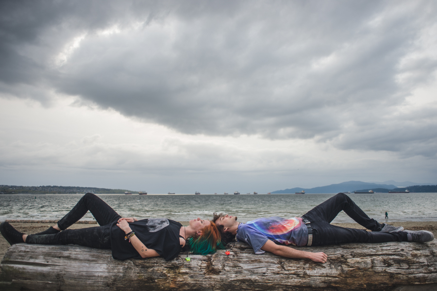 Two people lying with their heads near each other on a large shoreline log beneath a cloudy sky.