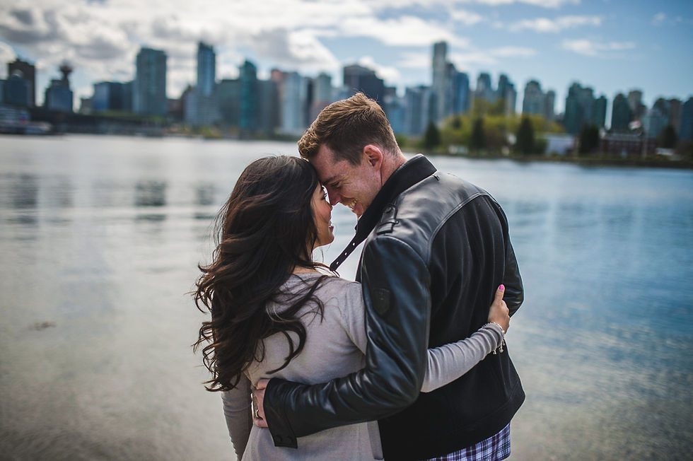 Couple touching foreheads beside the water with a city skyline behind them.