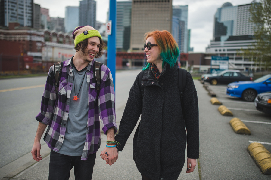 A smiling pair holding hands in a parking area with cars and tall buildings behind them.