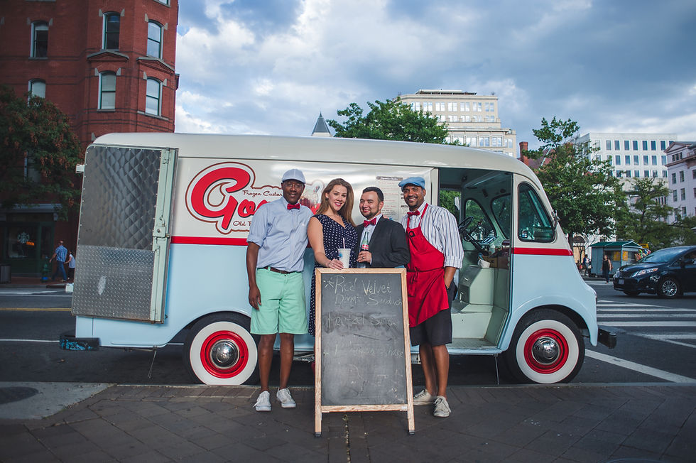 Four people stand beside a small white and red bus and a tall table.
