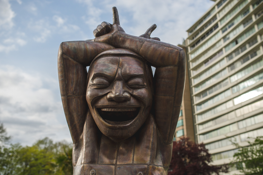 A bronze sculpture of a laughing figure with its arms raised above its head, beside a tall building.