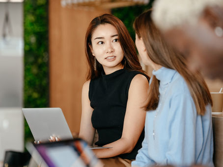 An Asian business woman wearing a black top is looking at her Asian female colleague during a conversation while sitting at a desk with a computer.