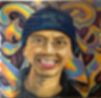 Mural-style portrait of a smiling young man wearing round glasses and a navy beanie reading "Trust Me I'm Greek," painted on brick against a colorful graffiti background.