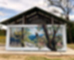 Mural on the gable end of a pavilion depicting regent honeyeaters in a bushland scene, one in flight over burnt tree trunks and one perched among gum blossoms.
