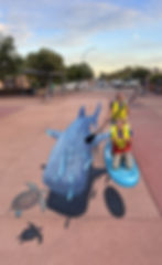 3D pavement art illusion of a large whale shark and sea turtles on a footpath, with two smiling women posing as if paddleboarding beside it.