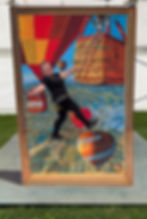 3D artwork of hot air balloons over a landscape, with a woman posing as if climbing a rope ladder toward the basket; a kingfisher perches on top. Signed "Jenny McCracken 2024."
