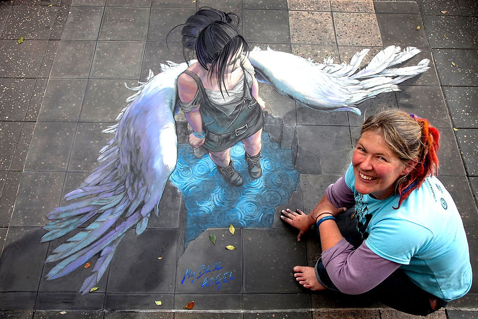 3D chalk pavement art titled "My Blue Angel," depicting a winged figure with dark hair standing in a rippling pool of water, viewed from above. The smiling artist lies beside her artwork on the pavement.
