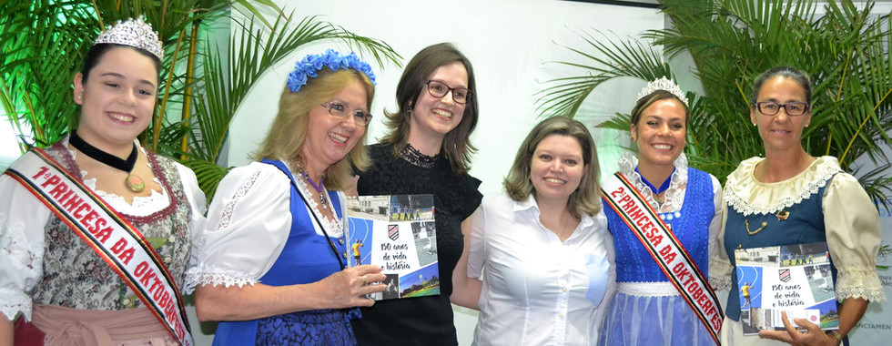 Autoras do livro Sogipa: 150 anos de vida e história, as sócias da Palavra Bordada, Denise Waskow e Carolina Rocha, posam para foto com a corte da Oktoberfest da Sogipa e integrantes do grupo dos Bávaros, vestidos com trajes típicos. Duas integrantes seguram exemplares da obra.