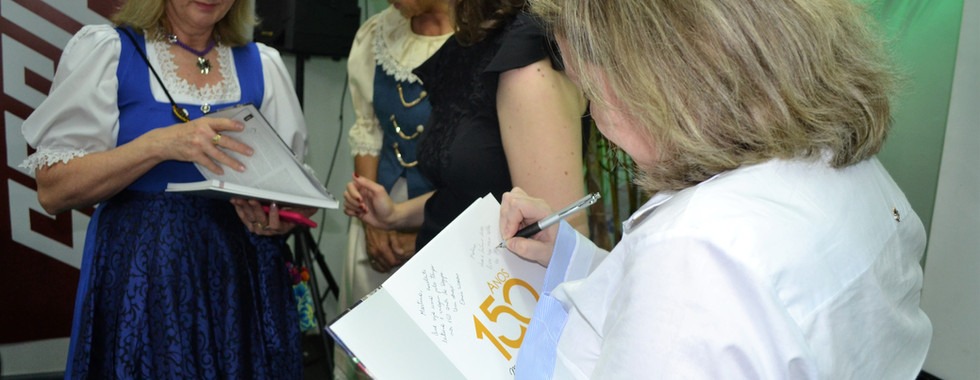 A sócia da Palavra Bordada, Carolina Rocha, e uma das autoras autografa um exemplar do livro Sogipa: 150 anos de vida e história. Ao fundo, a outra autora, a sócia da Palavra Bordada, Denise Waskow, conversa com duas integrantes do grupo dos Bávaros, a quem entrega um exemplar do livro.