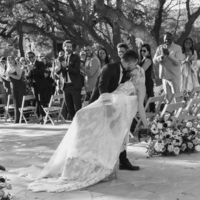 Black and white wedding photograph of a groom dipping his bride during their ceremony exit as he kisses her. The newlyweds are surrounded by cheering guests celebrating in the background.