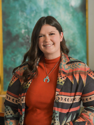 Woman in an orange and gray printed blazer and orange shirt stands and smiles for a headshot behind a teal picture