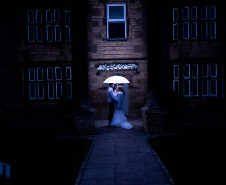 A bride and groom stand under a lit umbrella, framed by a dark stone building at dusk