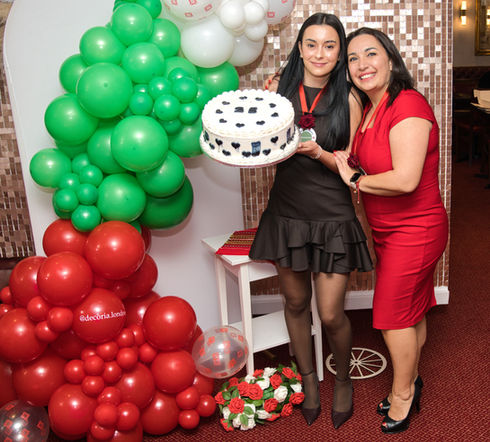 Two women holding a decorated cake beside green and red balloon display at a celebration event
