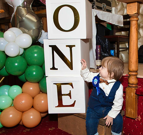 Toddler in blue dungarees beside large white ONE blocks at a first birthday party with green and white balloons