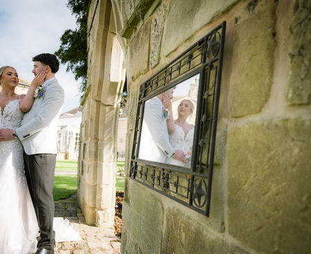 A bride and groom stand lovingly by an old stone wall, with their reflection visible in an ornate mirror