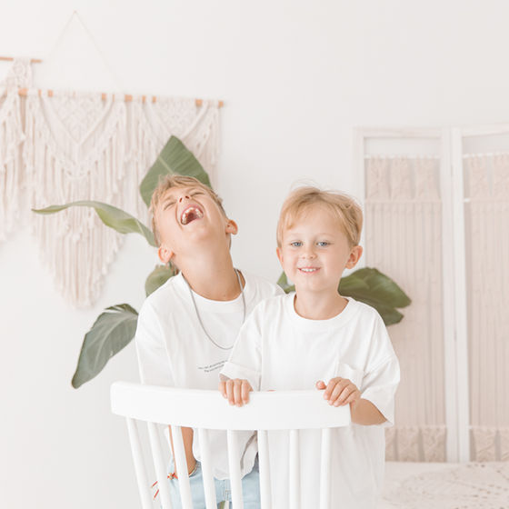 Kinderfotografie von Saskia Rachor: Zwei lachende Jungen hinter weißem Stuhl in einem hellen Raum.