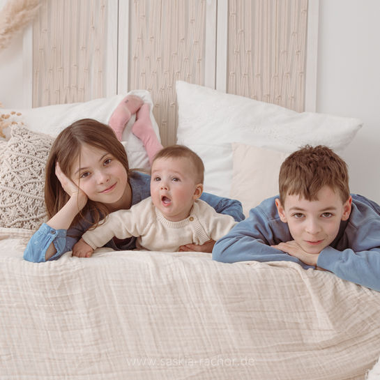 Drei lächelnde Geschwister, Baby in der Mitte, liegen auf hellem Bett.
