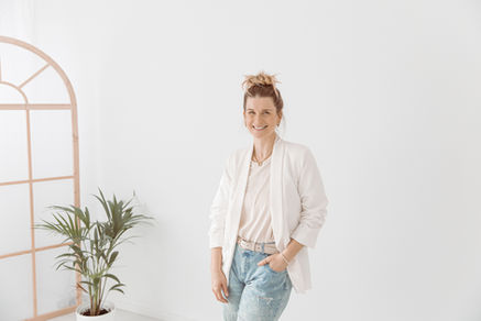 Lächelnde Frau in weißem Blazer und Jeans vor weißer Wand mit Pflanze.