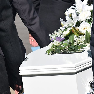 White coffin adorned with white lilies and greenery, carried by people in black suits