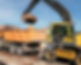 An excavator with a yellow cab loads dirt into an orange dump truck on a construction site