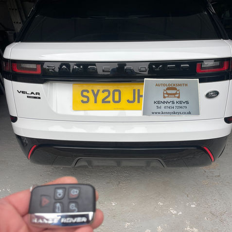A hand holding a Range Rover smart key fob behind a white Range Rover Velar with the licence plate SY20 JHH and a "Kenny's Keys" advertising card